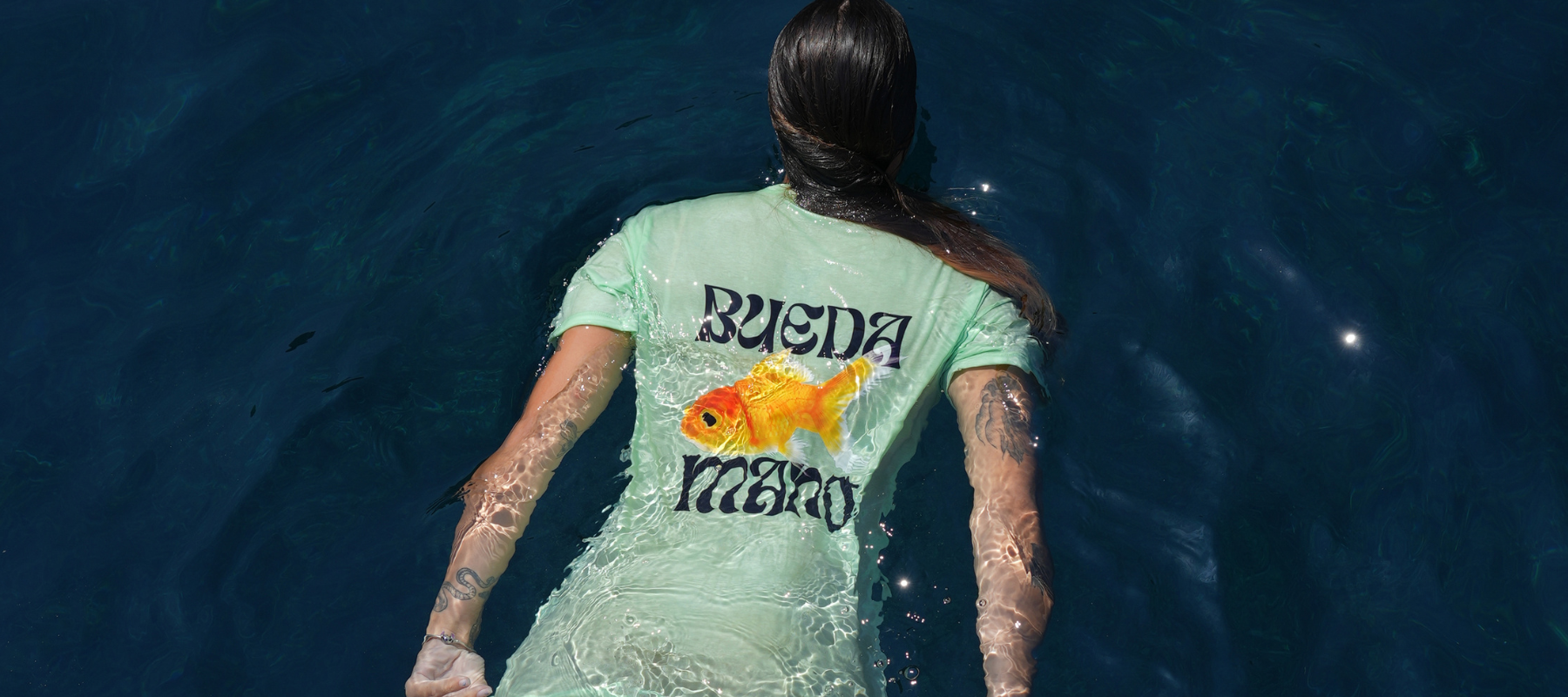 Person wearing a light green t-shirt THE KIKSON with text and a fish graphic, standing in water.